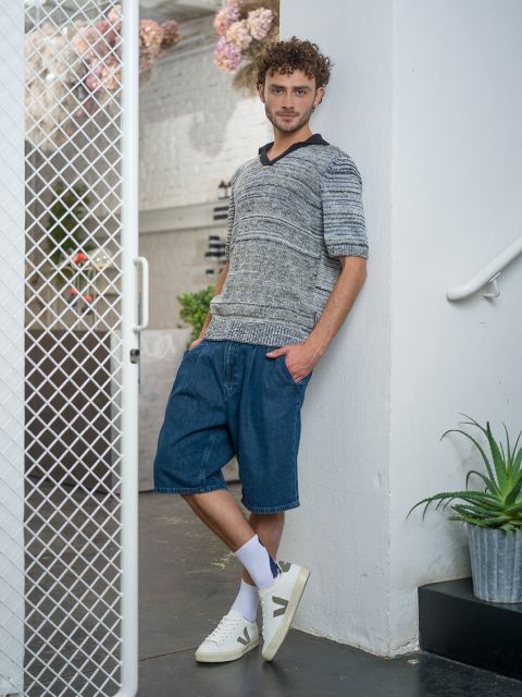 Ein Mann steht an einer weißen Wand, trägt einen grauen Strickpullover, Jeansshorts, weiße Socken und Turnschuhe, wobei er einen Fuß über den anderen schlägt.