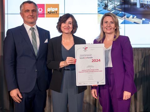 Drei Personen in Geschäftskleidung stehen zusammen, wobei die Frau in der Mitte eine Urkunde für 2024 in der Hand hält. Sie lächeln in die Kamera.