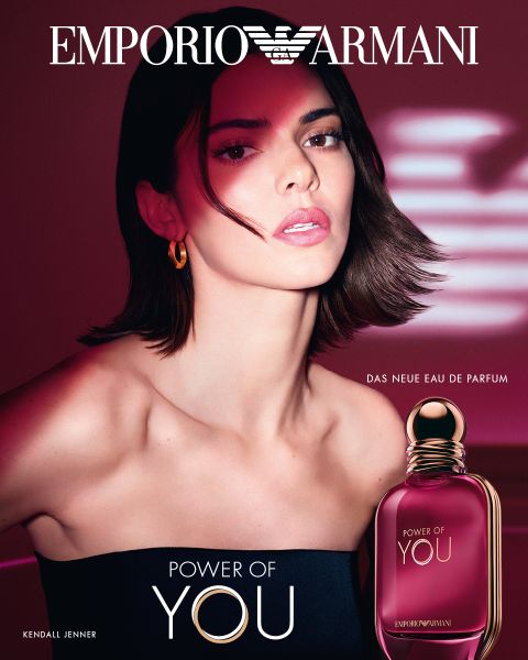 Eine Frau mit kurzen, dunklen Haaren und nackten Schultern posiert vor einem roten Hintergrund und wirbt für den Flakon des Parfüms "Power of You" von Emporio Armani im Vordergrund.