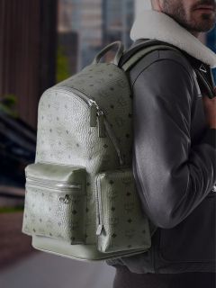 Eine Person in einer schwarzen Jacke trägt einen großen, strukturierten olivgrünen MCM-Rucksack mit Reißverschlusstaschen und steht im Freien in einer städtischen Umgebung.