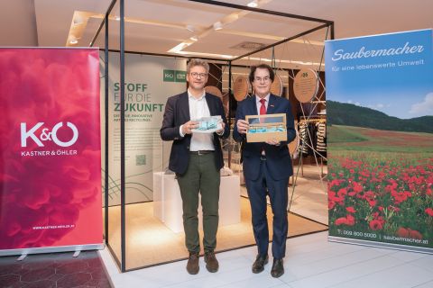 Zwei Männer in Anzügen stehen in einem modernen Geschäft und halten Kartons mit Gesichtsmasken in der Hand. Neben ihnen sind Werbebanner für "Kastner & Öhler" und "Saubermacher" zu sehen.