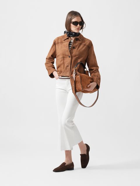 Frau in brauner Wildlederjacke, weißer Hose, dunklen Halbschuhen und Sonnenbrille, die eine passende braune Handtasche in der Hand hält, steht vor einem schlichten Hintergrund.