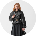 Frau mit langen roten Haaren, die eine schwarze Lederjacke mit Gürtel und einen dunklen Rock trägt, steht vor einem schlichten hellen Hintergrund und hält eine kleine Handtasche.