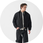 Eine Person, die eine schwarze Jacke über einem hellen Hemd und eine schwarze Hose trägt, steht vor einem schlichten, hellen Hintergrund.