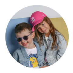 Zwei lächelnde Kinder in Jeansjacken; der Junge trägt eine Sonnenbrille, das Mädchen eine rosa Baseballmütze und lehnt ihren Kopf an seinen.