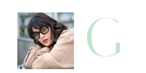 Frau mit dunklem Haar und Sonnenbrille, die einen beigen Mantel trägt und sich auf die Arme stützt, neben einem großen, hellgrünen Buchstaben "G" auf hellem Grund.