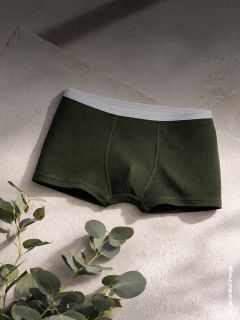 Dunkelgrüne Herren-Boxershorts mit weißem Bund sind auf einer strukturierten Oberfläche ausgestellt, mit Eukalyptusblättern in der Nähe in weichem, natürlichem Licht.