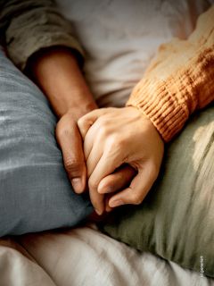 Zwei auf einem Bett liegende Personen, die sich an den Händen halten, wobei die eine einen orangefarbenen Ärmel und die andere einen grünen Ärmel trägt, um Trost und Nähe zu vermitteln.