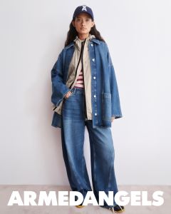 Eine Person in Jeans mit weitem Bein, Jeansjacke, gestreiftem Hemd, Mütze und Turnschuhen steht vor einer weißen Wand. Am unteren Rand steht "ARMEDANGELS" geschrieben.