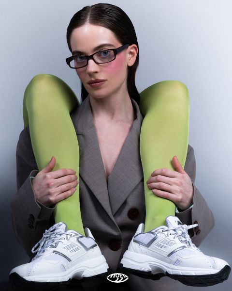 Eine Frau in einem grauen Blazer trägt eine Brille und weiße Turnschuhe und hält ihre Beine mit leuchtend grünen Strümpfen über die Schultern, vor einem schlichten Hintergrund.