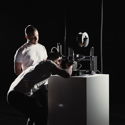 Zwei Personen bedienen und untersuchen einen 3D-Drucker auf einem weißen Sockel in einem dunklen Raum mit fokussierter Beleuchtung.