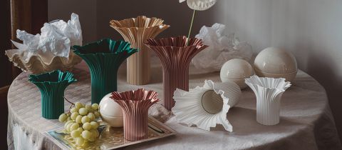 Auf einem Tisch stehen mehrere skulpturale Vasen in verschiedenen Farben und Größen, daneben Weintrauben, weiße Keramikkugeln und zerknülltes Papier.