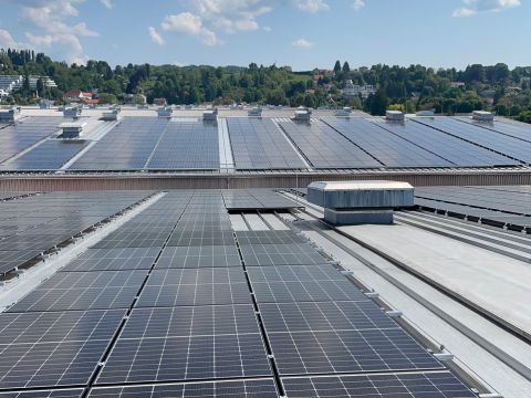 Reihen von Solarmodulen auf einem großen Dach unter einem blauen Himmel mit vereinzelten Wolken, mit Bäumen und Häusern im Hintergrund.