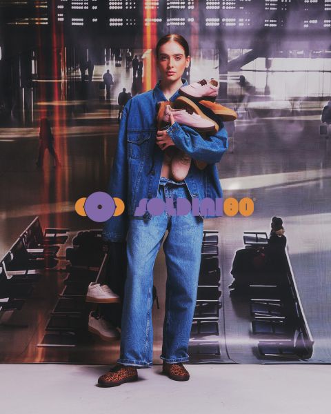 Eine Frau in Jeansjacke und Jeans steht in einem Flughafenterminal und hält mehrere Paar Turnschuhe in ihren Armen.