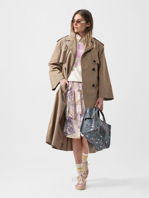 Frau in hellbraunem Trenchcoat, Sonnenbrille, pastellfarbenem grafischem Hemd, gemustertem Rock, gestreiften Socken und rosa Turnschuhen, die eine verzierte Jeanshandtasche in der Hand hält.