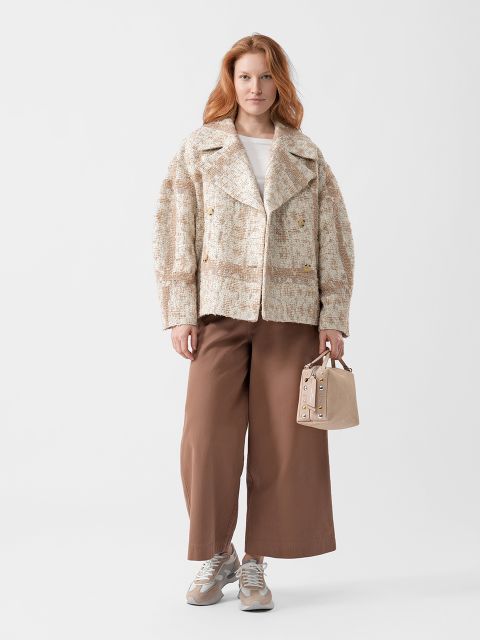 Frau mit langen roten Haaren trägt eine strukturierte beigefarbene Jacke, weite braune Hosen, Turnschuhe und hält eine kleine beigefarbene Handtasche. Sie steht vor einem weißen Hintergrund.