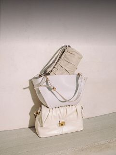 Drei elegante beigefarbene und weiße Handtaschen sind vor einer hellen Wand und einem Betonboden gestapelt und im natürlichen Sonnenlicht ordentlich angeordnet.