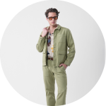 Mann in hellgrüner Jacke und Hose mit gemustertem Hemd, mit Sonnenbrille, steht vor einem schlichten weißen Hintergrund.