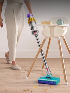 Eine Person benutzt einen schnurlosen Staubsauger, um einen Holzboden in der Nähe eines Hochstuhls und eines kleinen Tisches mit Geschirr von Krümeln zu befreien.