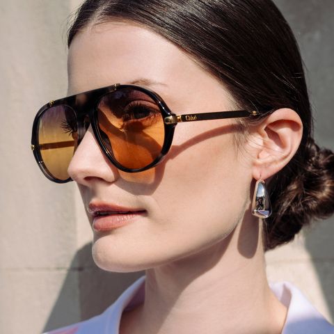 Frau mit dunklen Haaren in einem tiefen Dutt, die eine übergroße braune Sonnenbrille und silberne Ohrringe trägt und vor einer hellen Wand steht.