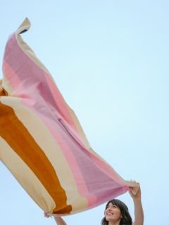 Eine Frau lächelt und hält eine große, bunt gestreifte Decke vor einem klaren blauen Himmel hoch.