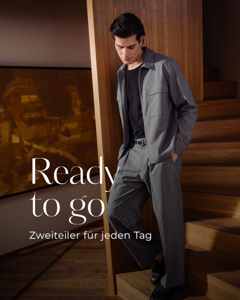 Mann im grauen Anzug mit schwarzem Hemd lehnt an einer Holztreppe. Der Text lautet "Ready to go. Zweiteiler für jeden Tag.".