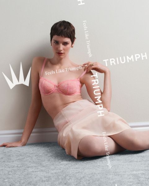 Eine Frau in einem rosa Spitzen-BH und einem durchsichtigen Rock sitzt auf einem Teppichboden vor einer Wand mit den Texten "Feels Like Triumph" und "TRIUMPH" um sie herum.