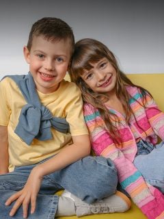 Ein lächelnder Junge und ein lächelndes Mädchen sitzen eng beieinander auf einer gelben Couch. Der Junge trägt ein gelbes Hemd, das Mädchen einen bunt gestreiften Pullover.