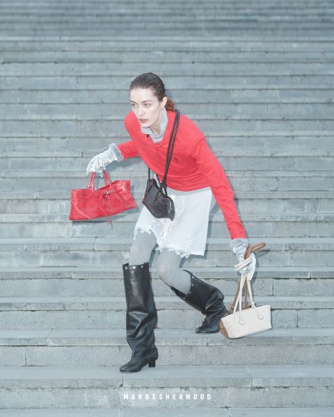 Eine Frau mit rotem Oberteil, grauen Handschuhen und schwarzen Stiefeln geht eine Steintreppe hinunter, in der Hand eine rote und eine beigefarbene Handtasche, darunter der Text "Margesherwood".