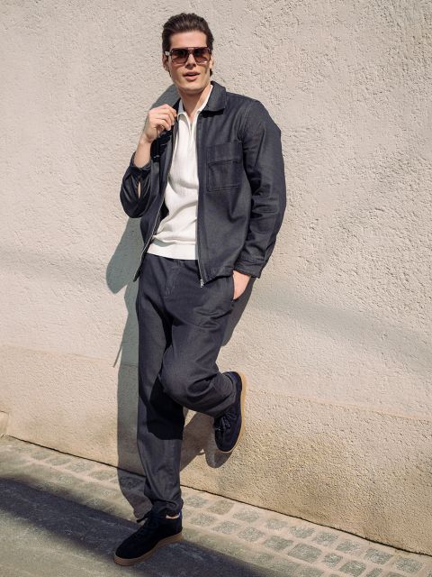 Ein Mann mit Sonnenbrille und dunklem Freizeitanzug lehnt an einer strukturierten Wand, eine Hand in der Tasche, während das Sonnenlicht Schatten wirft.