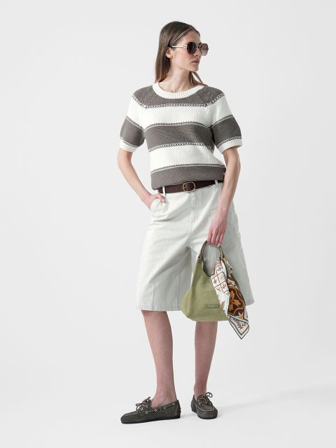 Frau mit Sonnenbrille, die einen gestreiften kurzärmeligen Pullover, weiße knielange Shorts und Halbschuhe trägt und eine grüne Handtasche mit einem Schal in der Hand hält.