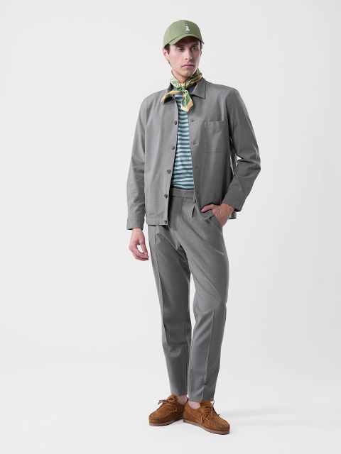 Mann in grauem Anzug, gestreiftem Hemd, grüner Mütze, gemustertem Halstuch und braunen Schuhen vor einem schlichten weißen Hintergrund.