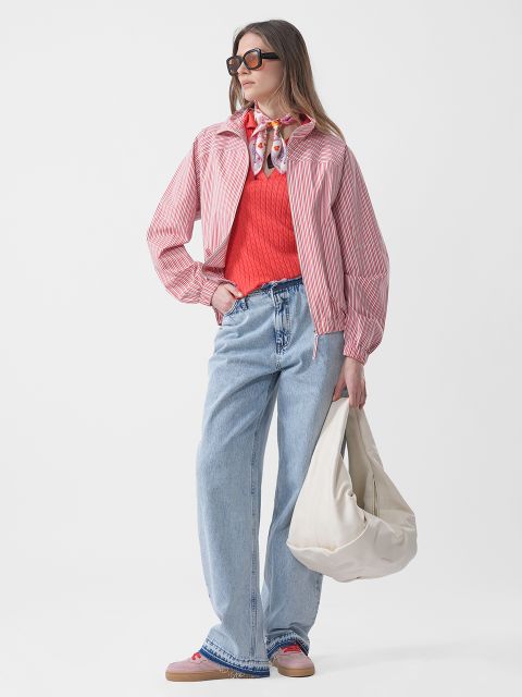 Frau mit Sonnenbrille trägt eine rosa gestreifte Jacke, einen roten Pullover, blaue Jeans und einen Schal und hält eine große weiße Tasche vor einem schlichten weißen Hintergrund.