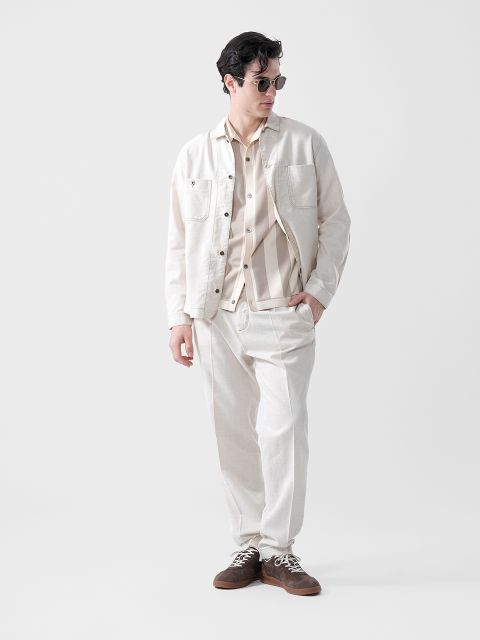 Ein Mann mit Sonnenbrille, hellbeiger Jacke, gestreiftem Hemd und passender Hose steht mit den Händen in den Taschen vor einem schlichten weißen Hintergrund.