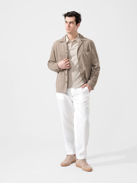 Mann in beiger Jacke, hellem Polohemd, weißer Hose und hellbraunen Turnschuhen vor weißem Hintergrund, eine Hand in der Tasche.