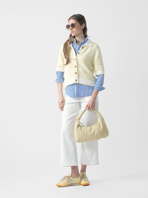 Frau mit Sonnenbrille, gestreiftem blauen Hemd, gestreifter Strickjacke, weißer Hose und gelben Schuhen hält eine hellgelbe Handtasche und steht vor einem weißen Hintergrund.