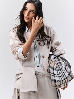 Frau in hellbeiger Kleidung mit einer karierten Tasche, die zur Seite schaut und ihr Haar vor einem schlichten Hintergrund berührt.