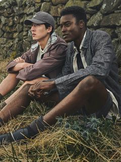 Zwei Männer sitzen in legerer Outdoor-Kleidung auf trockenem Gras an einer Steinmauer und schauen nachdenklich in die Ferne.