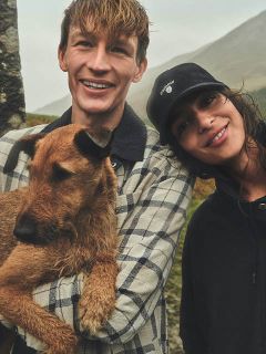 Ein lächelnder Mann hält einen braunen Hund, während er neben einer lächelnden Frau mit schwarzer Mütze steht, im Freien an einem bewölkten Tag.