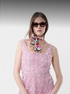 Frau in einem rosa Kleid mit Blumenmuster, mit Sonnenbrille und einem bunten Schal um den Hals, vor einem neutralen Hintergrund.
