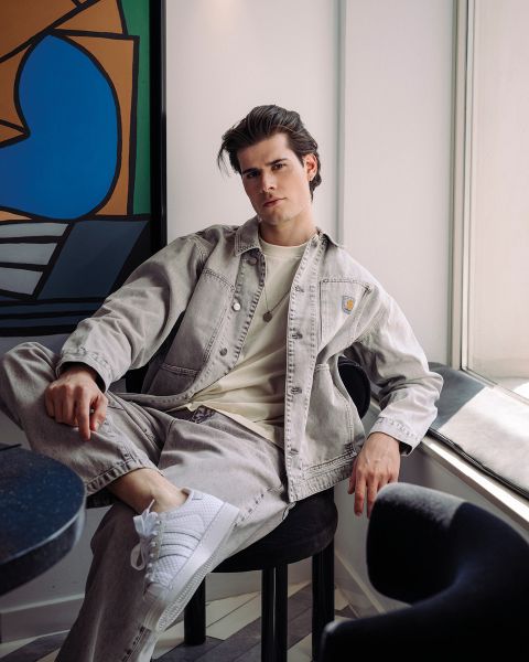 Ein junger Mann in einer hellen Jeansjacke und weißen Turnschuhen sitzt entspannt auf einem Stuhl am Fenster in einem modernen Raum mit abstrakter Kunst an der Wand.