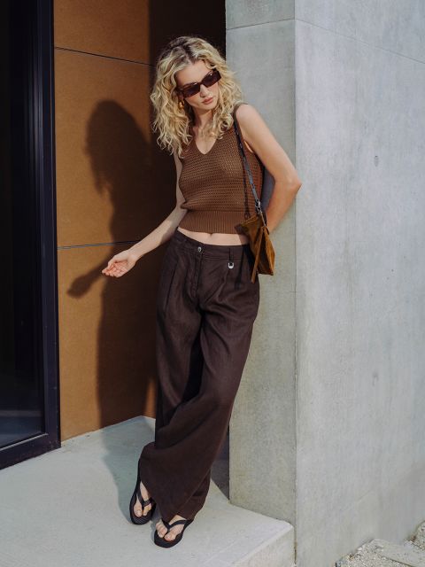Eine Frau mit lockigem blondem Haar trägt eine Sonnenbrille, ein braunes ärmelloses Oberteil, eine braune Hose mit weitem Bein und Sandalen und steht vor einem modernen Gebäude.