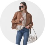 Frau mit Sonnenbrille, brauner Fransenjacke, cremefarbenem Pullover, blauen Jeans und weißer Handtasche vor einem hellen Hintergrund.