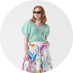 Frau mit Sonnenbrille, die eine mintgrüne Strickjacke, einen geblümten Rock und einen Schal trägt und eine rosa Handtasche in der Hand hält, steht vor einem weißen Hintergrund.