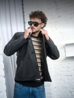 Ein Mann mit Sonnenbrille, schwarzer Lederjacke, gestreiftem Pullover und Jeans steht vor einer weißen Backsteinmauer und rückt selbstbewusst seinen Kragen zurecht.