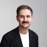 Lächelnder Mann mit kurzen braunen Haaren und einem Schnurrbart, der einen schwarzen Blazer über einem weißen Hemd trägt, posiert vor einem schlichten hellgrauen Hintergrund.
