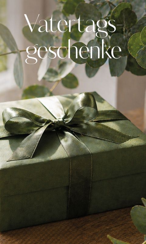 Grüne Geschenkschachtel mit einem glänzenden Band auf einem Holztisch, umgeben von grünen Blättern. Text: "Vatertagsgeschenke" (Vatertagsgeschenke) oben.