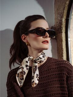 Frau mit dunklem Haar in einem Pferdeschwanz, die eine schwarze Sonnenbrille, goldene Ohrringe, einen braunen Pullover und einen Schal mit Leopardenmuster trägt und zur Seite schaut.