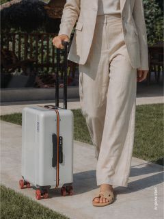 Eine Person in einem beigen Anzug geht nach draußen und zieht einen weißen Hartschalenkoffer mit roten Rädern und schwarzem Griff über einen gepflasterten Weg.