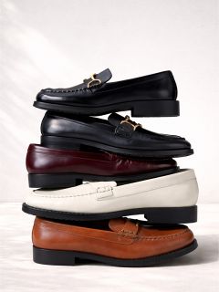 Ein Stapel von vier Lederslippern in Schwarz, Burgunderrot, Weiß und Braun, die vertikal vor einem hellen Hintergrund angeordnet sind.